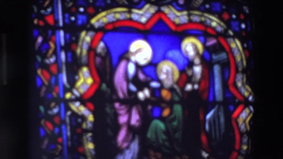 Soft close-up from 1969 Paris footage shows a stained glass religious scene with haloed figures, saturated blue glass, and dark surrounding church interior.