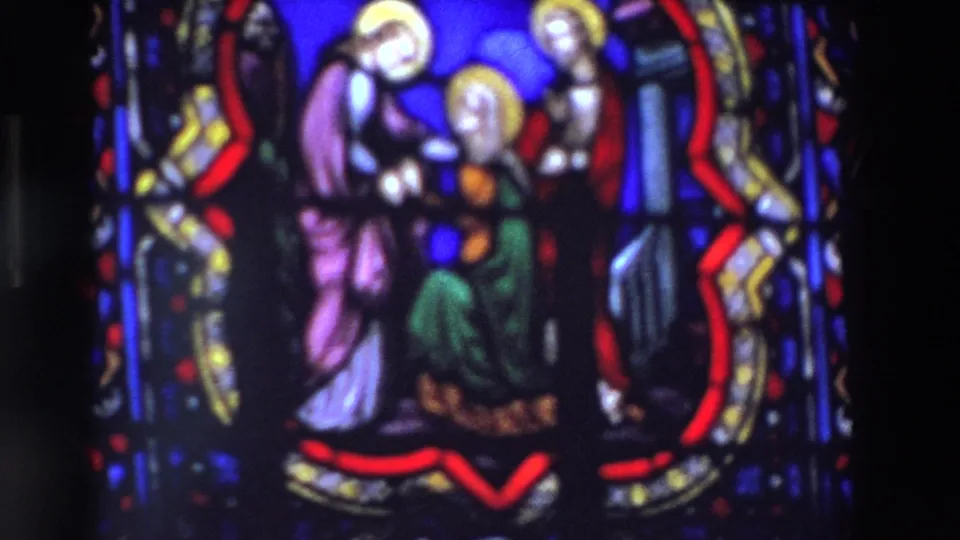 Close visitor-shot 8mm view of stained glass religious figures in a Paris church or cathedral setting, with strong blue glass and red decorative borders.