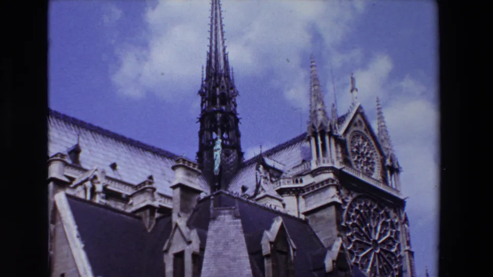 1969 Paris 8mm frame showing Notre-Dame Cathedral exterior with roofline, Gothic spire, rose window, and pale clouds against blue sky.
