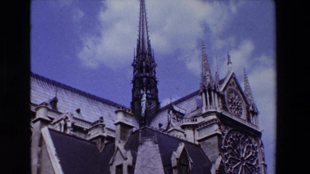 1969 Paris home-movie frame showing Notre Dame Cathedral's spire, Gothic roofline, stone details, and rose window against a blue sky with clouds.