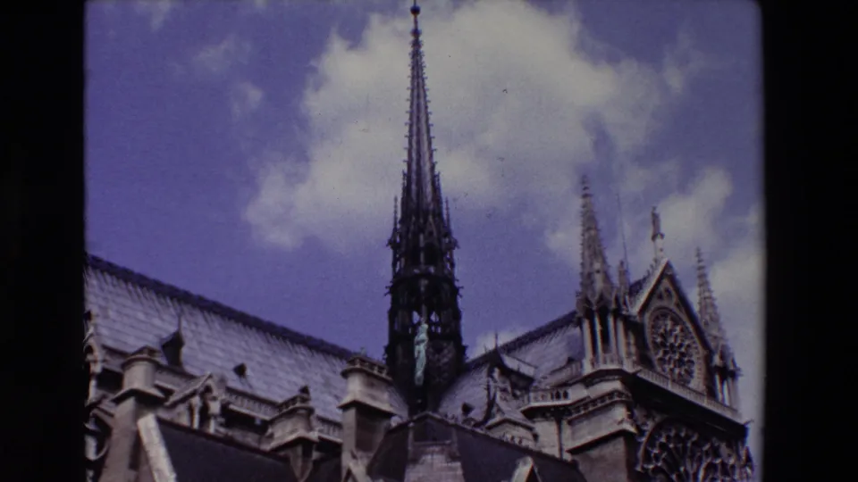 1969 Paris home-movie view looking up at Notre Dame Cathedral's spire, decorative stonework, roofline, and rose window beneath scattered clouds.