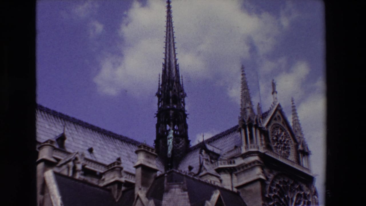 Paris, France 1969 8mm frame showing a Notre-Dame Cathedral spire rising above steep rooflines and carved stone ornament under a cloudy sky.