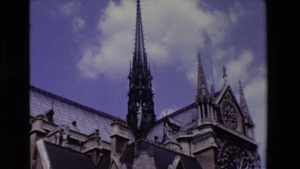 Paris, France 1969 8mm frame showing a Notre-Dame Cathedral spire rising above steep rooflines and carved stone ornament under a cloudy sky.