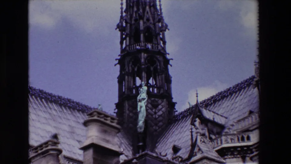 Paris, France 1969 8mm frame showing an angelic statue mounted near a dark Gothic spire above the Notre-Dame Cathedral roofline.