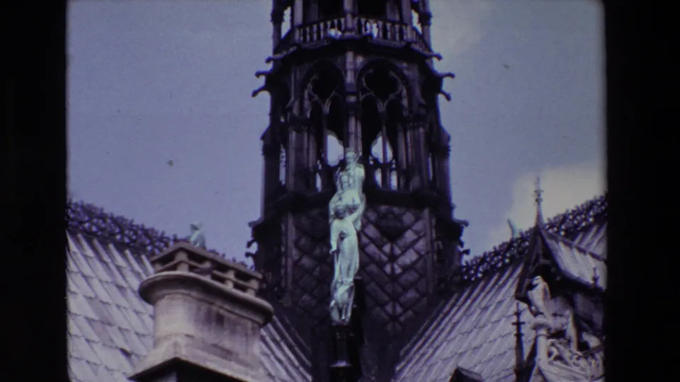 Paris, France 1969 8mm frame with a closer visitor-shot view of an angelic figure attached to a Notre-Dame Cathedral spire, framed by roof slopes and stone ornament.
