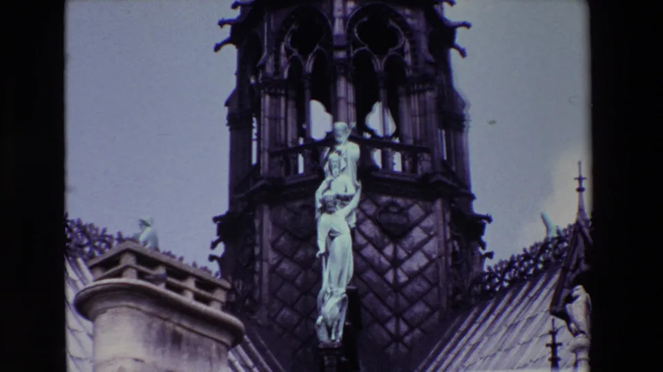 1969 Paris 8mm frame showing a Gothic cathedral spire with tracery, roof ornament, and a pale religious statue group viewed from below.