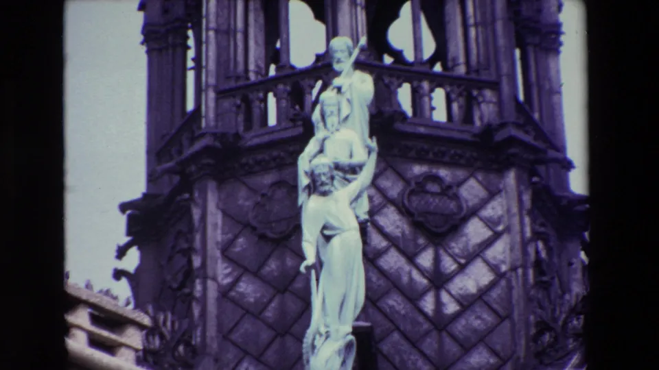 1969 Paris 8mm frame with the camera closer to a pale religious sculpture set against dark patterned stone and open Gothic tracery.