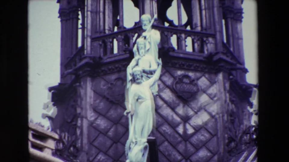 1969 Paris 8mm frame centering the pale religious statue group with patterned masonry, spire details, and roof ornament around it.