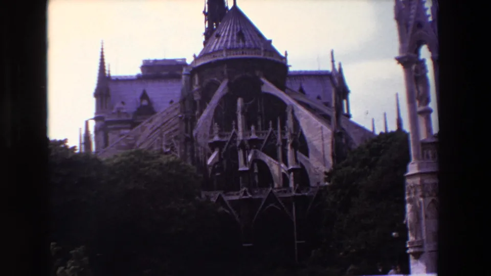 1969 Paris 8mm frame showing a wider visitor-shot view of a Gothic cathedral exterior, with flying buttresses, spires, roof detail, and trees across the lower frame.