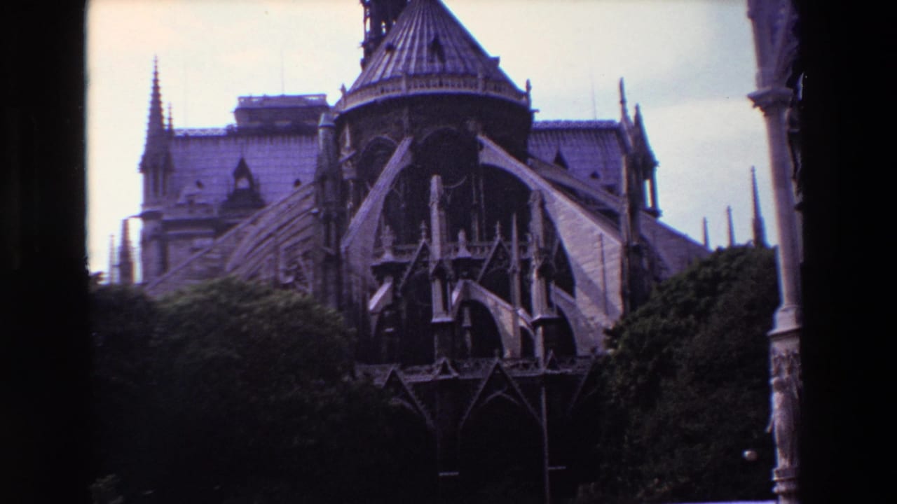 1969 8mm frame of Notre-Dame Cathedral in Paris showing the rear elevation, flying buttresses, pointed arches, roofline, spires, trees, and black film borders.