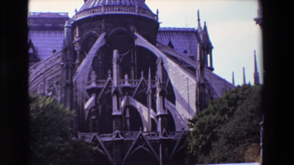 1969 8mm frame from Paris with a closer view of Notre-Dame Cathedral's flying buttresses, pointed arches, roofline, spires, and dense greenery at the lower edge.