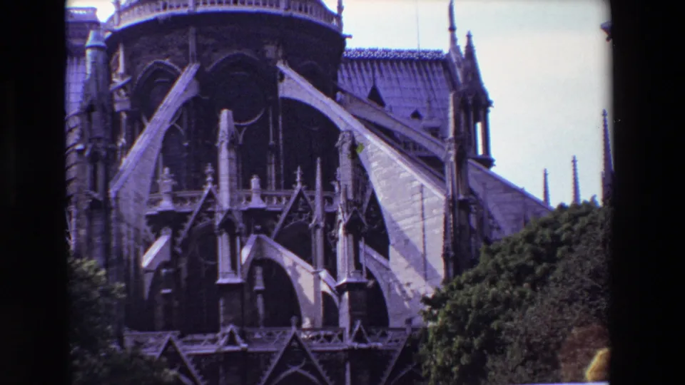 1969 Paris frame showing Notre-Dame Cathedral's buttressed rear elevation, dark arched openings, decorative stonework, roof structure, spires, and trees along the foreground.