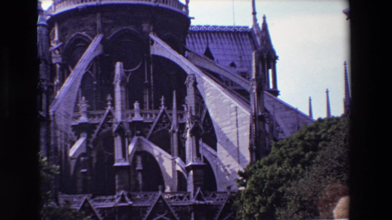 1969 Paris 8mm frame showing Notre-Dame Cathedral exterior with flying buttresses, pointed arches, roof tiles, pale sky, and greenery at the lower edge.