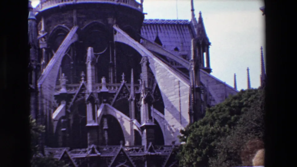 1969 Paris 8mm frame showing Notre-Dame Cathedral exterior with flying buttresses, pointed arches, roof tiles, pale sky, and greenery at the lower edge.