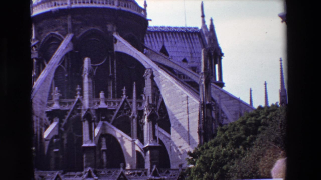 1969 Paris 8mm frame of Notre-Dame Cathedral with large exterior buttresses, shadowed arch openings, tiled roof sections, and green shrubbery in the foreground.