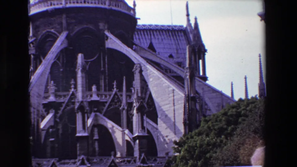 1969 Paris 8mm frame of Notre-Dame Cathedral with large exterior buttresses, shadowed arch openings, tiled roof sections, and green shrubbery in the foreground.