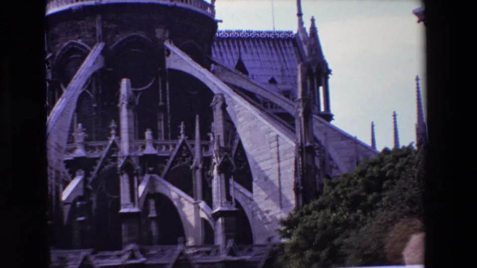 1969 Paris 8mm frame holding on Notre-Dame Cathedral's exterior buttresses, pointed arch forms, stone ornament, roof edge, and bushes across the bottom of the frame.