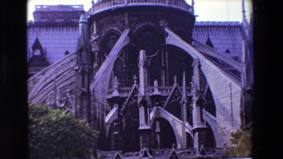 1969 Paris 8mm frame showing Notre-Dame Cathedral's Gothic exterior with flying buttresses, decorative pinnacles, roof surfaces, pale sky, and greenery in the lower foreground.