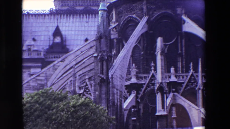 1969 Paris 8mm frame with a closer handheld view of Notre-Dame's exterior buttresses, roof plane, vertical stonework, and leafy foreground.