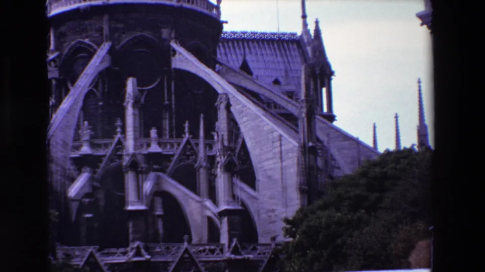 1969 Paris 8mm frame showing Notre-Dame Cathedral's Gothic side elevation with multiple buttresses, small pinnacles, dark recesses, and trees below.