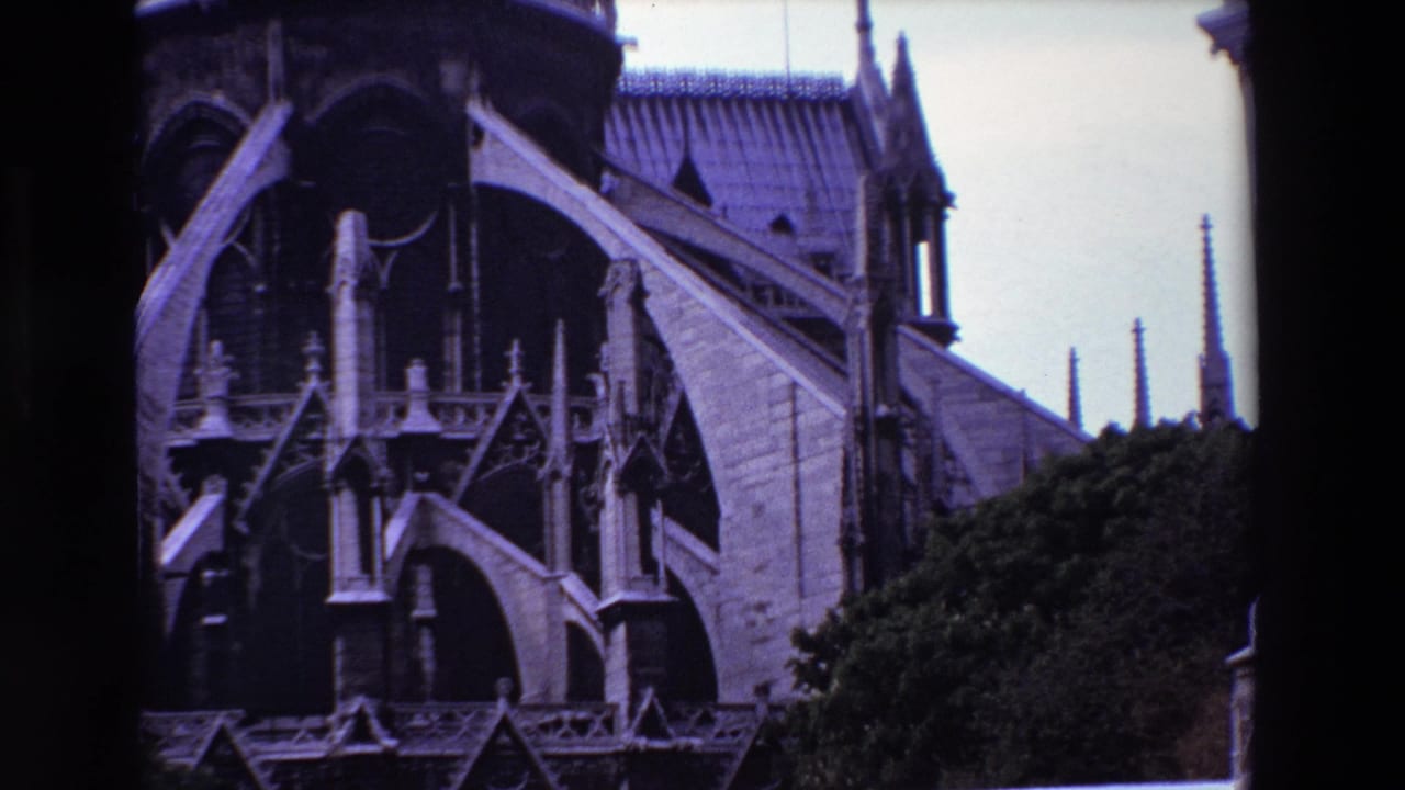 1969 Paris home-movie frame showing Notre-Dame Cathedral's upper exterior, with pale stone flying buttresses, dark Gothic arches, roof tiles, spires, greenery, and the edge of the 8mm scan.