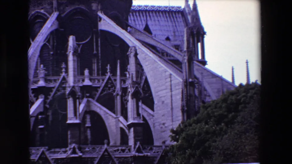 1969 Paris frame of Notre-Dame Cathedral's Gothic exterior, with large stone buttresses crossing the facade, pointed arches in shadow, a tiled roof, slender spires, and trees at the lower right.