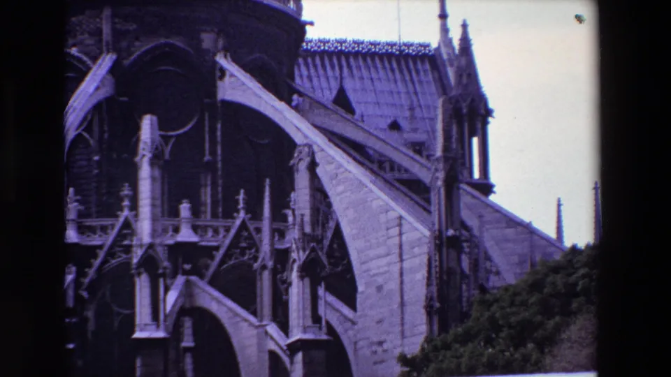 1969 Paris 8mm still showing Notre-Dame Cathedral's upper facade through dark scan borders, including flying buttresses, carved stone details, pointed arches, roof tiles, spires, and surrounding greenery.