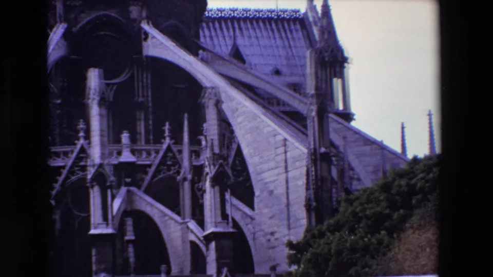 1969 Paris home-movie frame capturing Notre-Dame Cathedral from a slightly shifted angle, with a prominent flying buttress, dark arch openings, roof tiles, vertical spires, cloudy sky, and trees below.