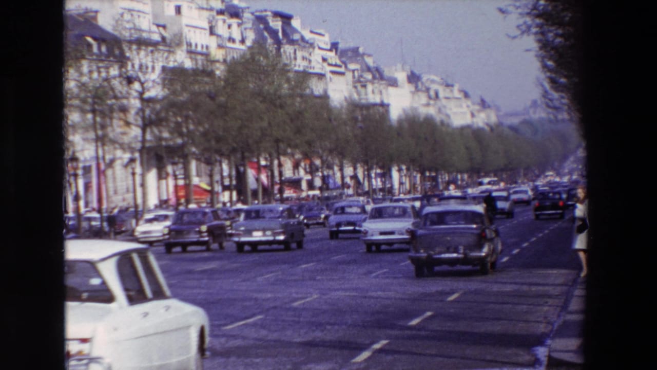 1969 Paris home-movie frame showing vintage cars moving along a broad tree-lined street bordered by apartment buildings and dark film edges.