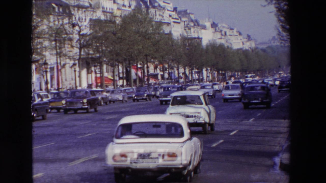 1969 Paris street traffic with a white compact car in the foreground, older cars across several lanes, trees, sidewalks, and apartment facades in the background.