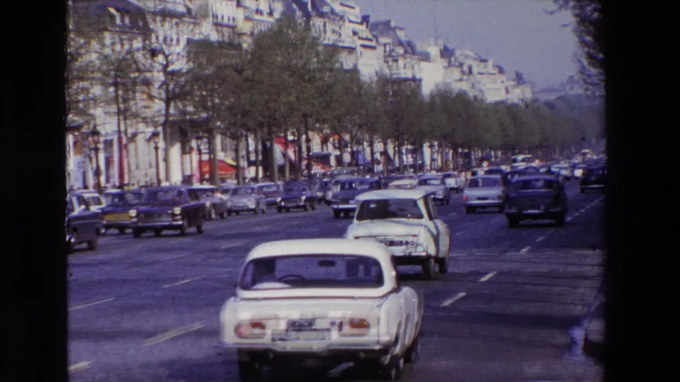 1969 Paris street traffic with a white compact car in the foreground, older cars across several lanes, trees, sidewalks, and apartment facades in the background.