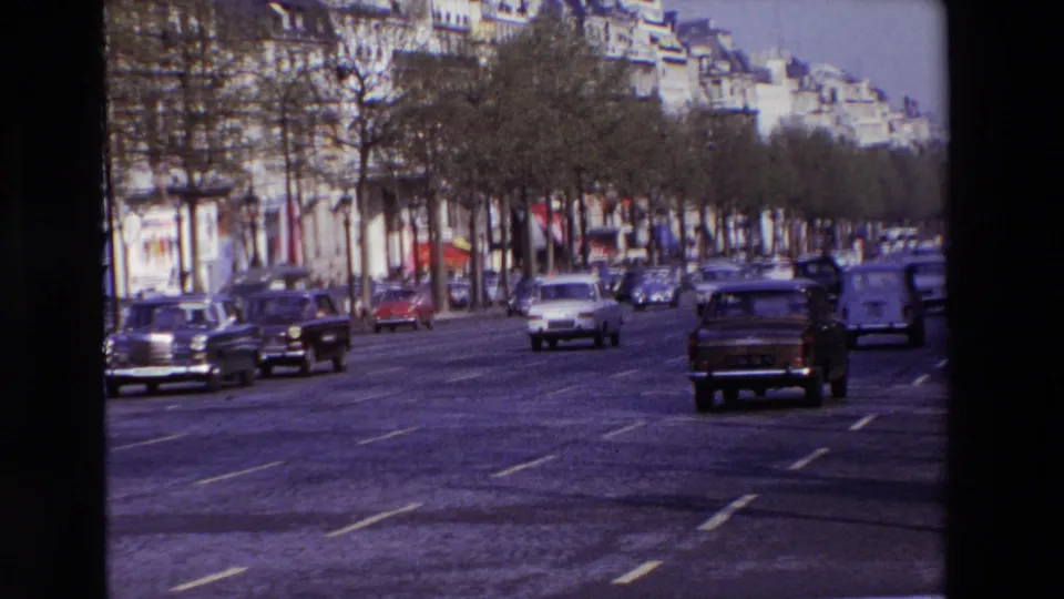 Vehicle-shot 1969 Paris footage showing several vintage cars spread across a wide road, with trees, storefront-level facades, and soft 8mm color visible.