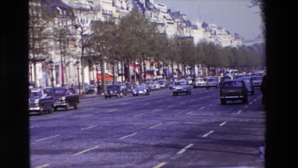 1969 Paris home-movie frame with cars receding along a broad tree-lined street, apartment buildings to the left, and dark 8mm gate edges around the frame.
