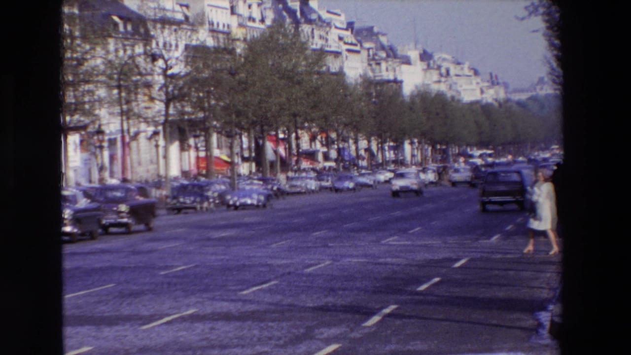 1969 Paris street scene showing a broad tree-lined boulevard, mid-century cars, apartment facades, and a woman in a light coat or dress crossing from the right edge.