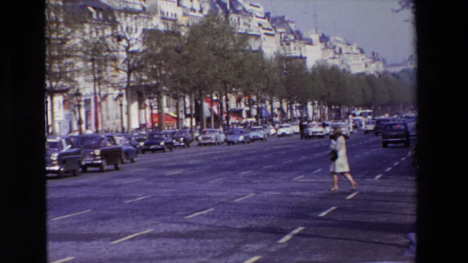 1969 Paris boulevard view with a woman in light clothing crossing multiple lanes while mid-century cars travel along a tree-lined street.