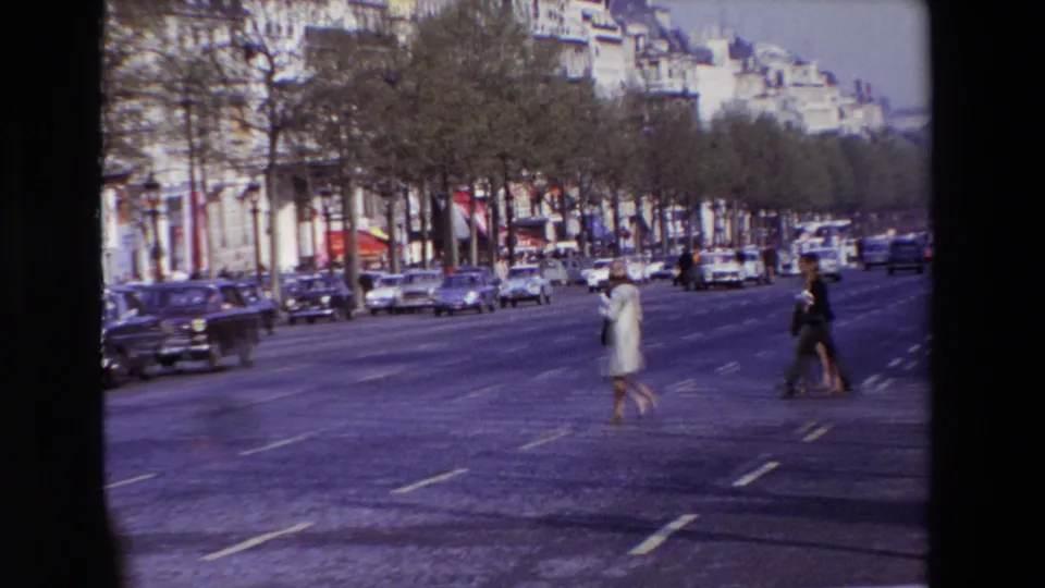 1969 Paris street footage showing a woman in light clothing and another pedestrian crossing a broad boulevard with cars, trees, and storefronts in the background.