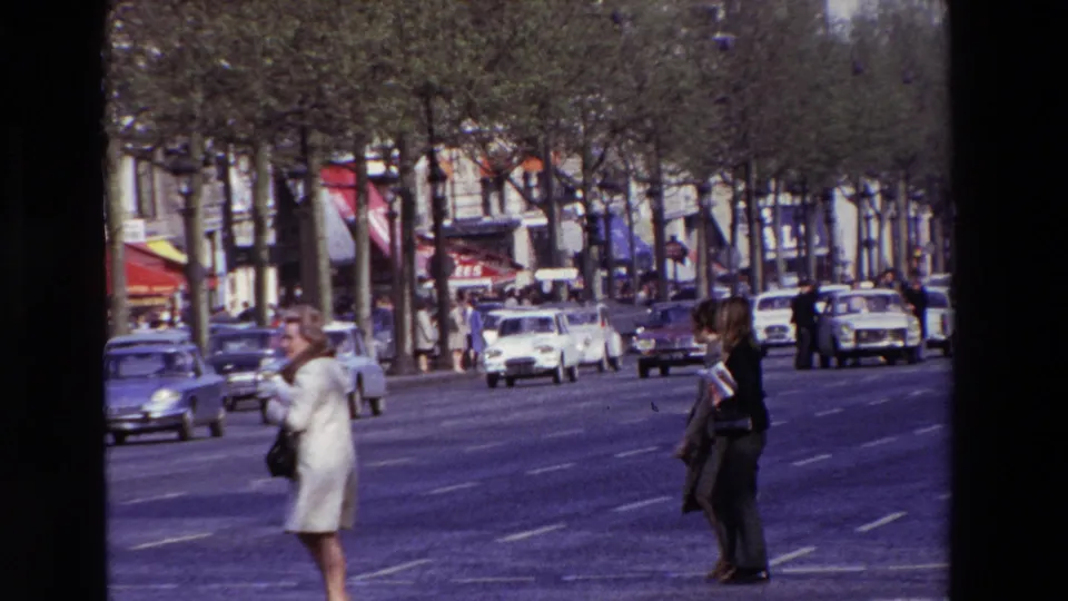 Closer 1969 Paris boulevard frame with a woman in light clothing and another pedestrian crossing in front of traffic, trees, awnings, and mid-century cars.