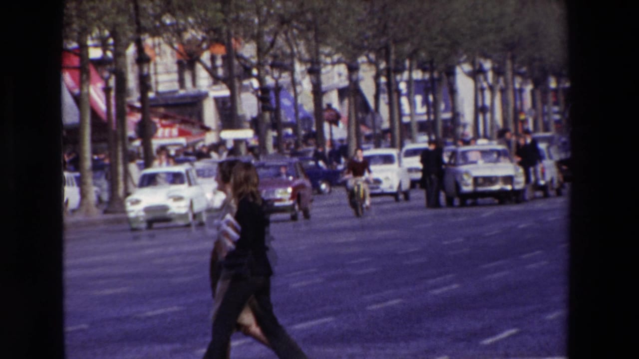 In 1969 Paris, a woman in dark clothing crosses a broad city street while compact cars, pedestrians, trees, and storefront awnings fill the background.