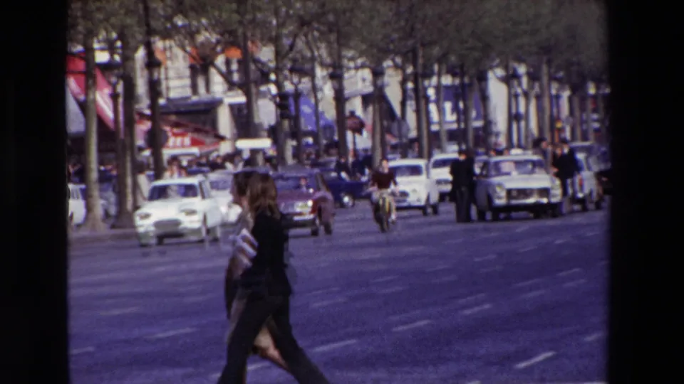 In 1969 Paris, a woman in dark clothing crosses a broad city street while compact cars, pedestrians, trees, and storefront awnings fill the background.
