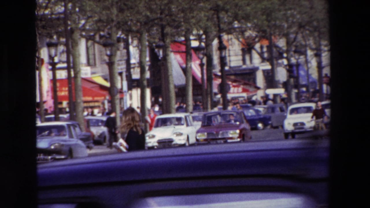 The moving-vehicle view shows 1969 Paris street traffic, a pedestrian near the roadway, compact cars, tree trunks, storefronts, and the dark edge of the car interior in the foreground.