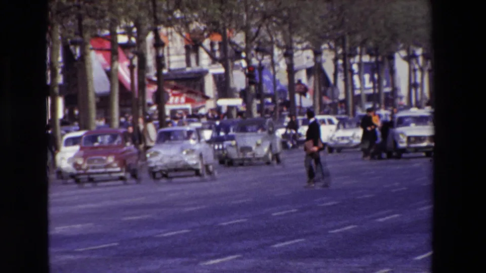 A 1969 Paris boulevard scene shows multiple compact cars moving along the road, pedestrians crossing or waiting, rows of trees, and commercial storefronts beyond the traffic.
