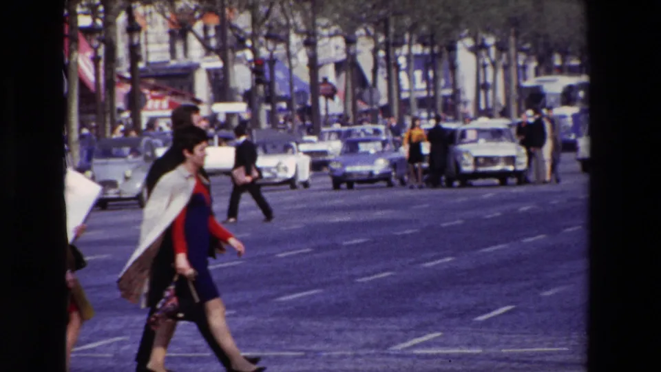 In 1969 Paris, pedestrians in period clothing cross a broad boulevard in front of compact cars, with tree trunks, lane markings, and storefronts visible behind them.