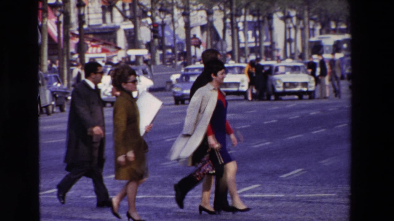 Paris, France in 1969, with suited men and women in coats crossing a broad boulevard amid period cars and storefronts.
