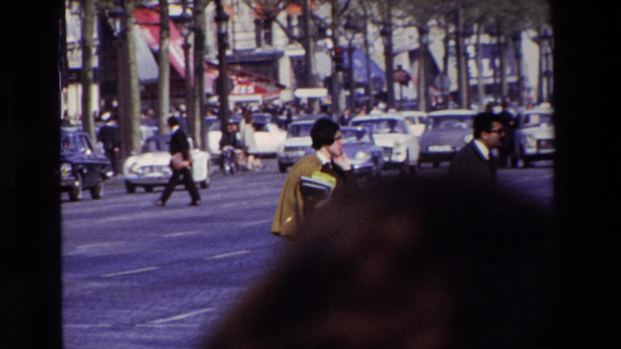A woman carrying papers crosses or walks along a wide Paris boulevard in 1969, with cars, storefronts, and pedestrians behind her.