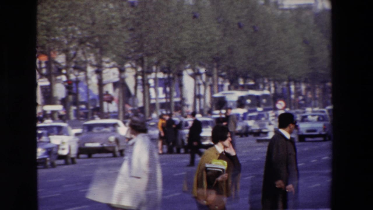 Pedestrians pass along the edge of a boulevard with traffic and trees in the background.
