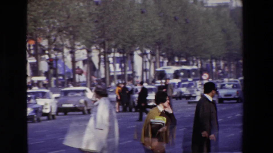 Paris street pedestrians in 1969 pass near a broad boulevard, with a suited man, a woman carrying papers, cars, and bare trees visible.
