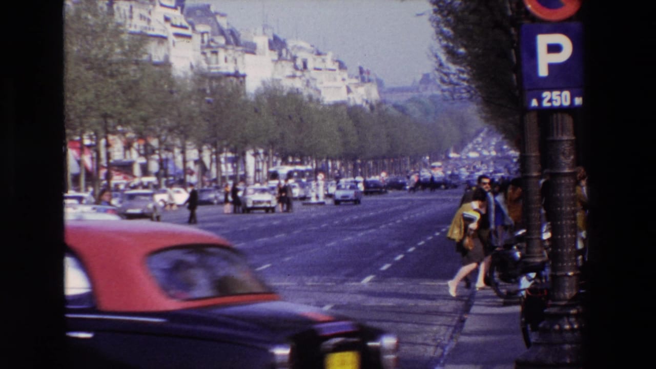 1969 Paris home-movie frame showing a broad avenue near the Arc de Triomphe, with classic cars, pedestrians, apartment buildings, and a parking sign visible.