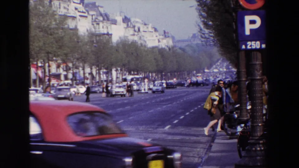 1969 Paris home-movie frame showing a broad avenue near the Arc de Triomphe, with classic cars, pedestrians, apartment buildings, and a parking sign visible.
