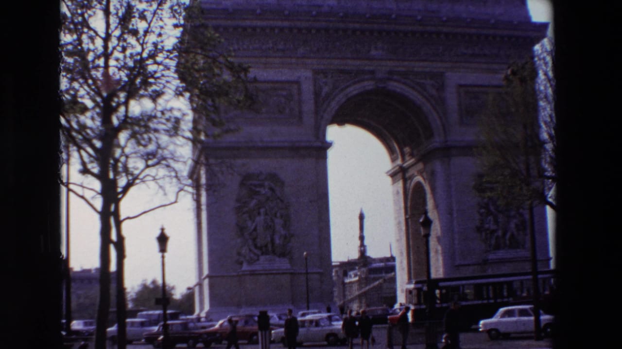 1969 Paris frame of the Arc de Triomphe from street level, with pedestrians, classic cars, bare trees, and lamp posts around the monument.
