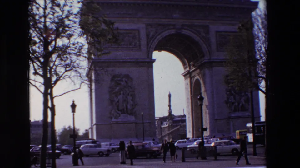 1969 Paris home-movie still showing the Arc de Triomphe partly framed by trees, with classic cars and pedestrians moving near the base of the monument.